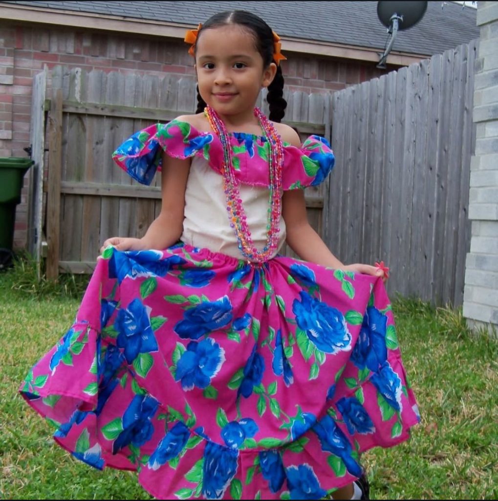 Aura Chuck Hernandez as a young girl, wearing a traditional Mexican dress and pig-tail braids.
